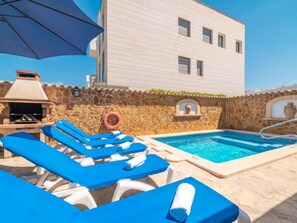 Zona de piscina amb gandules blaves, una barbacoa i una paret de pedra.