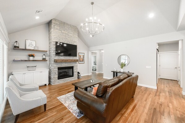 A spacious living room with a stone fireplace, a leather sofa, a striped armchair, and a wooden coffee table.