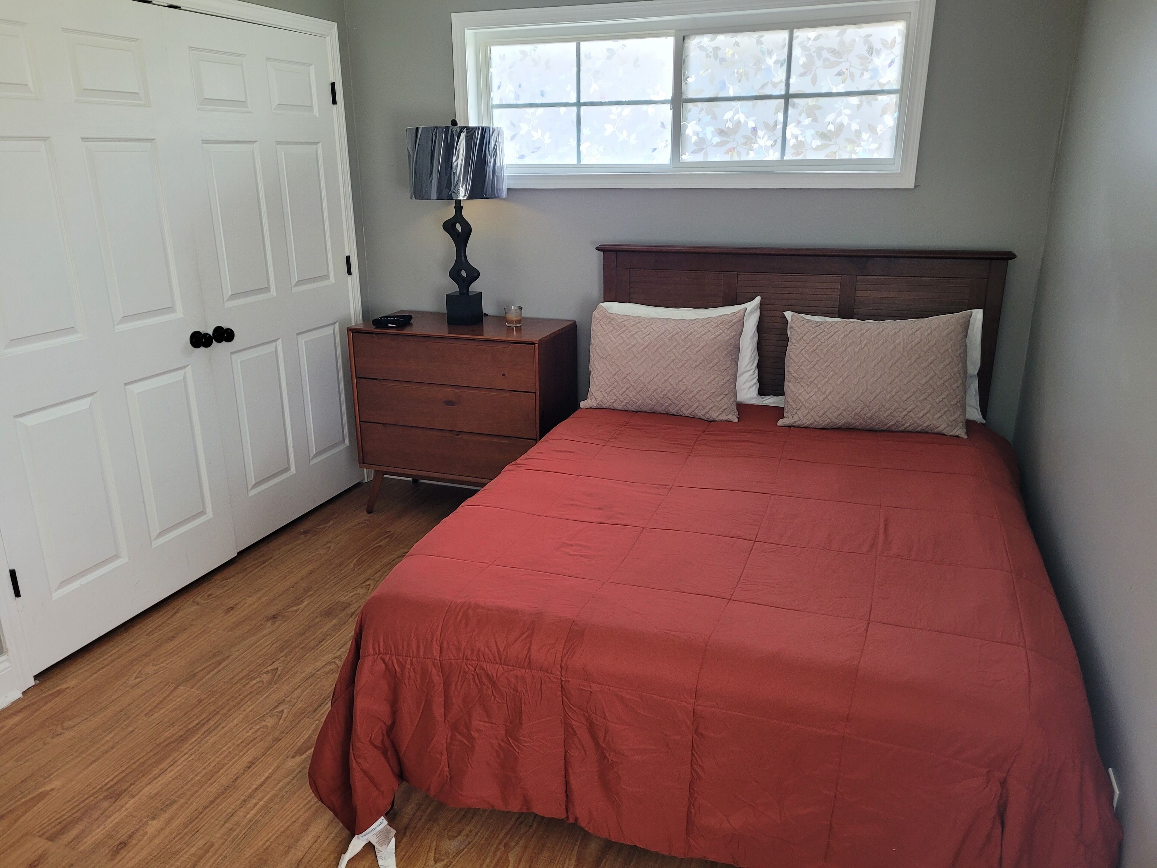 A bedroom with a wooden dresser, a red bedspread, two pillows, a nightstand with a lamp, and a window with floral blinds.