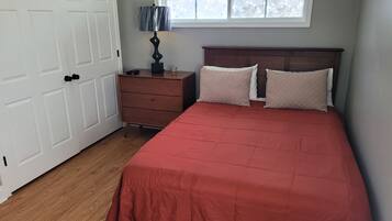 A bedroom with a wooden dresser, a red bedspread, two pillows, a nightstand with a lamp, and a window with floral blinds.