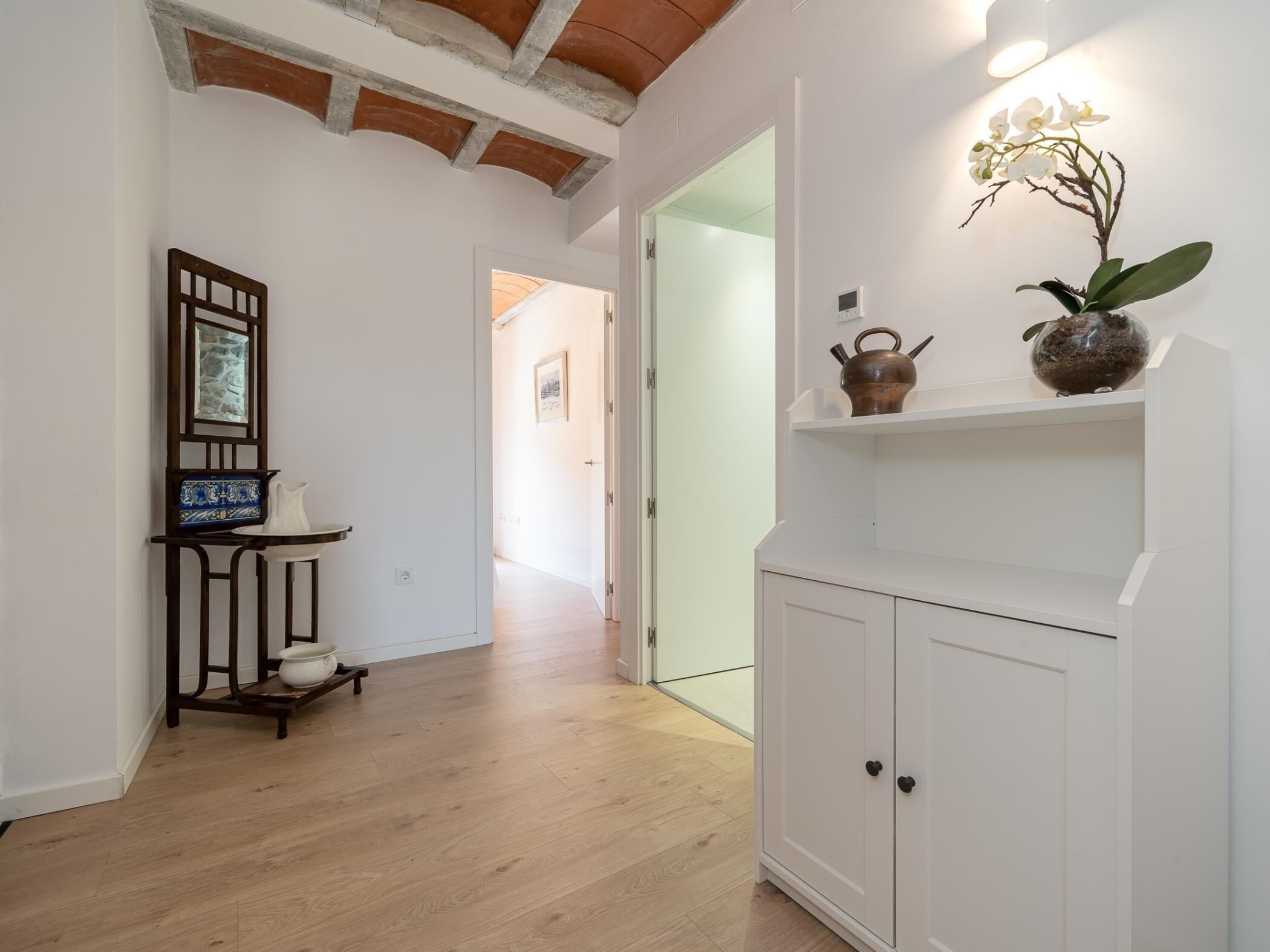 A hallway with wooden flooring, a white cabinet with black handles, a shelf with decorative items, and a framed picture on a stand.