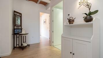 A hallway with wooden flooring, a white cabinet with black handles, a shelf with decorative items, and a framed picture on a stand.