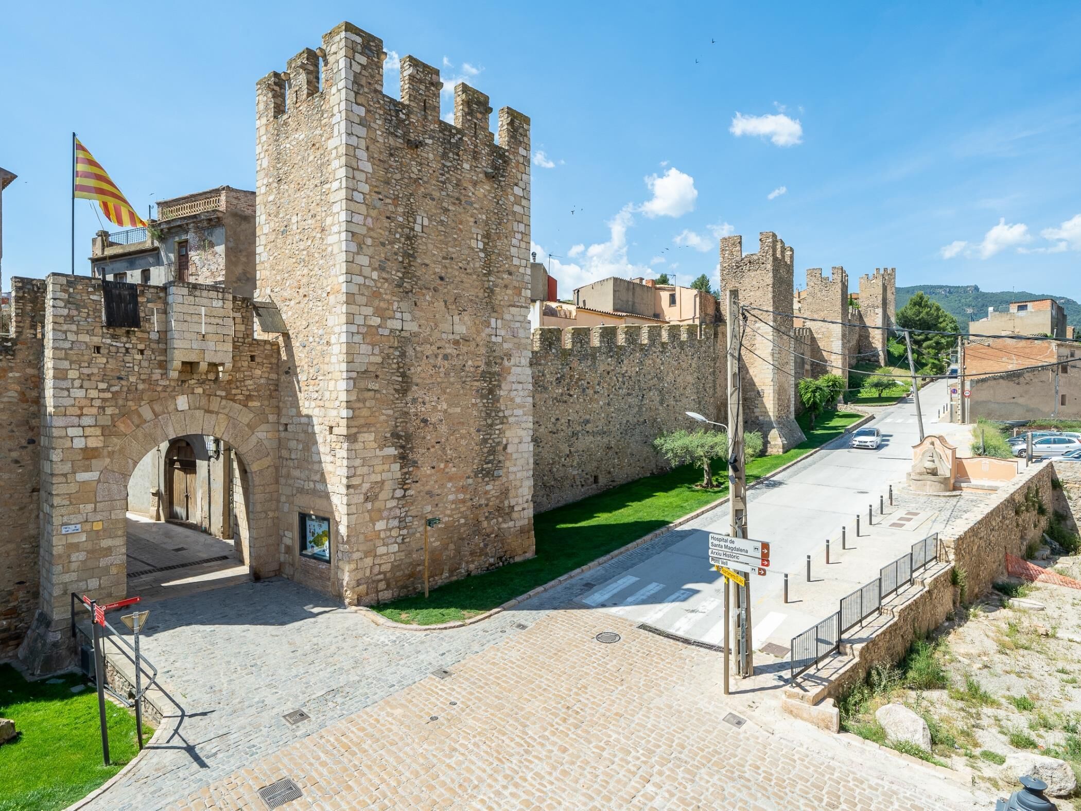 Un château historique en pierre, doté d’une grande entrée en arc de cercle, d’un drapeau et d’une rue pavée avec des panneaux de signalisation.