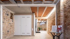 A hallway with stone walls, a tiled floor, and a white locker. There is a table with a sign and a potted plant in the distance.