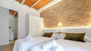 A hotel room with two beds, a wooden ceiling, a brick wall, and a nightstand with a lamp.