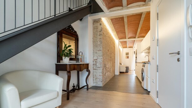 Un intérieur moderne avec un canapé blanc, une table en bois agrémentée d’une plante, et un coin cuisine doté de meubles blancs et d’un dosseret carrelé.