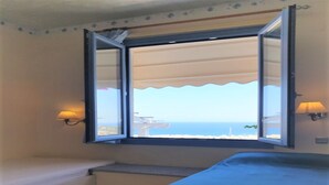 Ein Zimmer mit einem Bett, einer Bank, einem Fenster mit freiem Meerblick und zwei Wandleuchten.