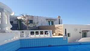 Ein Schwimmbecken mit blauer Fliesenbordur, einem weißen Geländer und einem Gebäude mit steinverkleideter Fassade und grünen Türen.