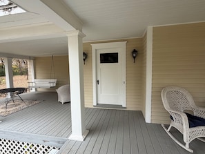 A covered porch with a white door, two wall-mounted lights, a wicker rocking chair, and a bench.
