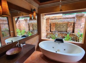 Um banheiro com uma banheira oval grande, uma pia redonda e um espelho. Há uma janela com vista para uma parede de tijolos e plantas.