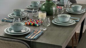 Een eettafel gedekt met tealkleurige borden, glazen en bestek, een decoratieve vaas en een kleurrijke pompon.