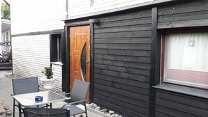 Ein kleiner Sitzbereich im Freien mit Tisch und Stühlen, eine Holztür und ein Fenster mit Vorhang.