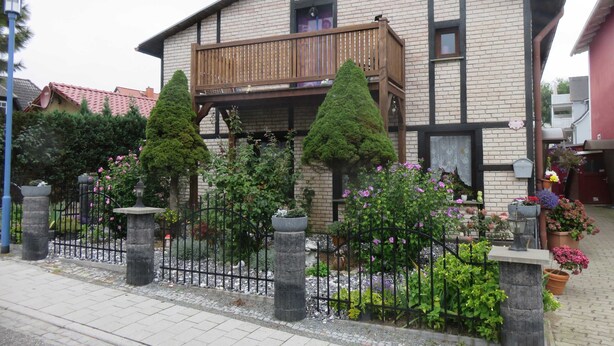 Ein zweistöckiges Haus mit einem Balkon, umgeben von einem Steinzaun und verschiedenen Pflanzen.