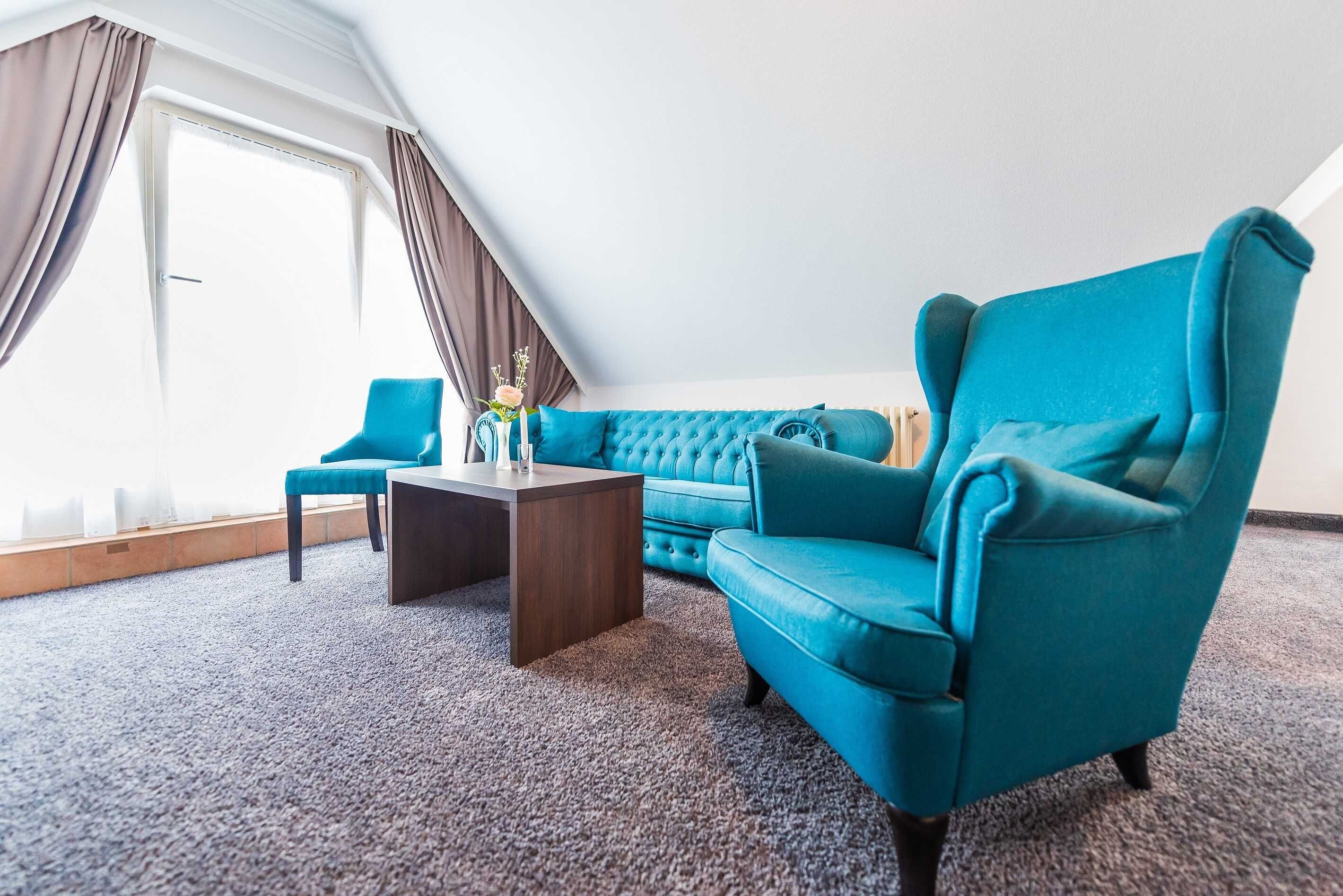 A room with a teal armchair, a blue sofa, a wooden coffee table, and a view of the outdoors through a large window with curtains.
