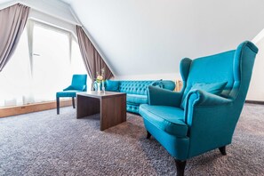 Une pièce comprenant un fauteuil turquoise, un canapé bleu, une table basse en bois et une vue sur l’extérieur grâce à une grande fenêtre agrémentée de rideaux.