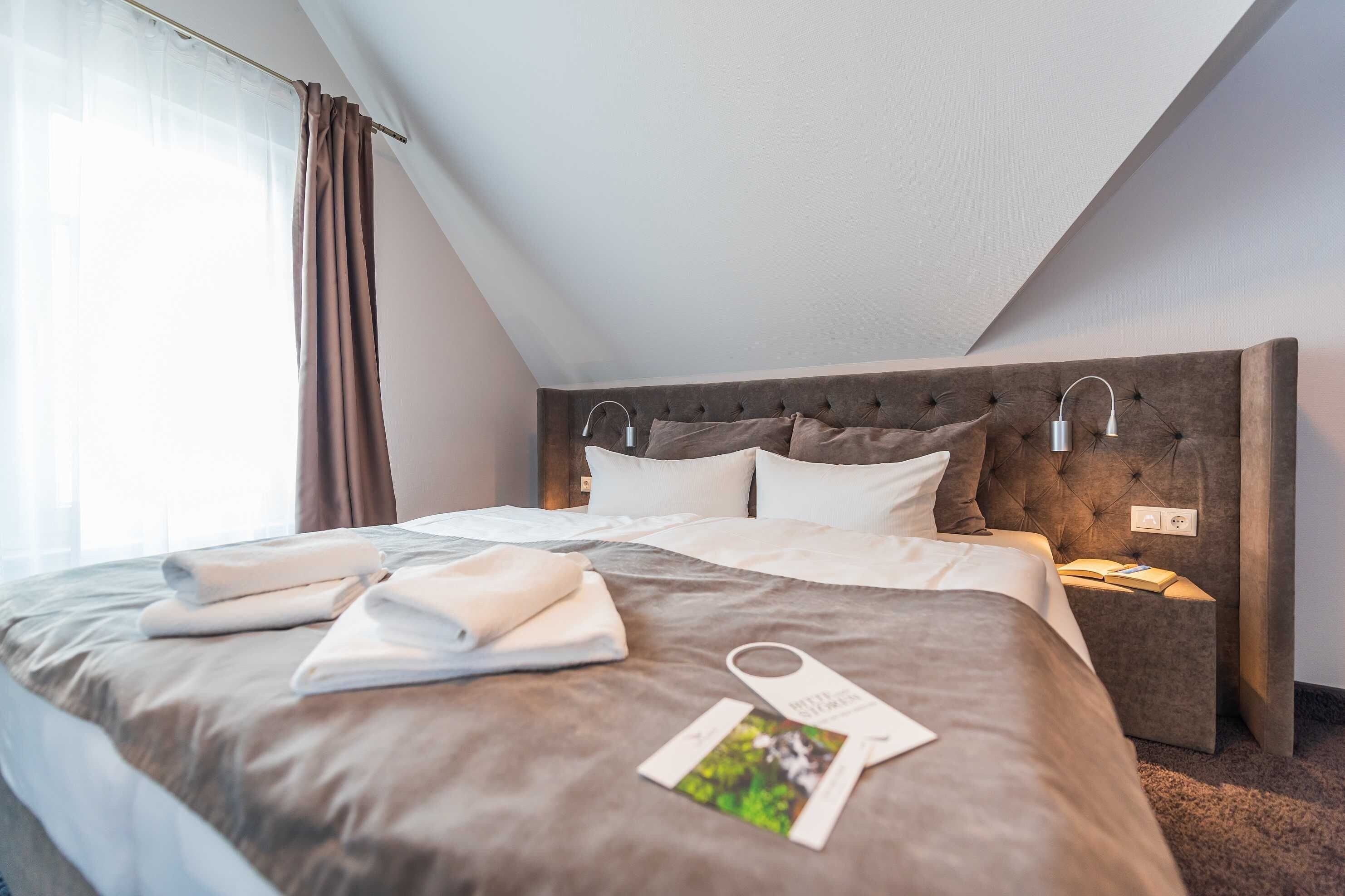 Ein ordentlich bezogenes Bett mit Kissen und einem gefalteten Handtuch, ein Nachttisch mit einer Lampe und ein Fenster mit Vorhängen.