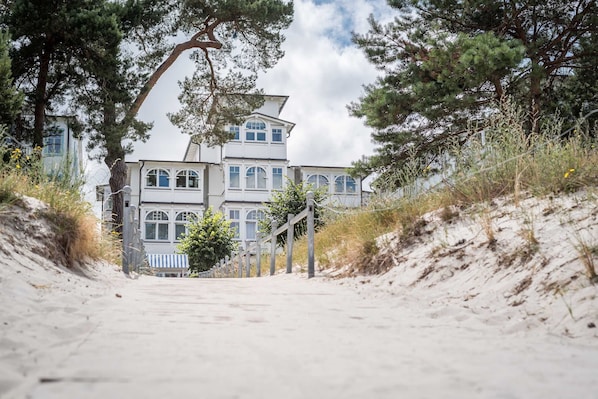A coastal building with a symmetrical facade, a staircase leading up to the entrance, and a sandy path.