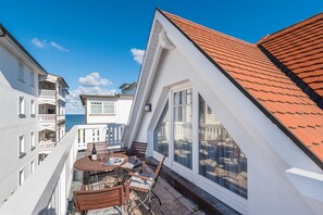 A balcony with a table and chairs, a view of the sea, and a building with a red roof.