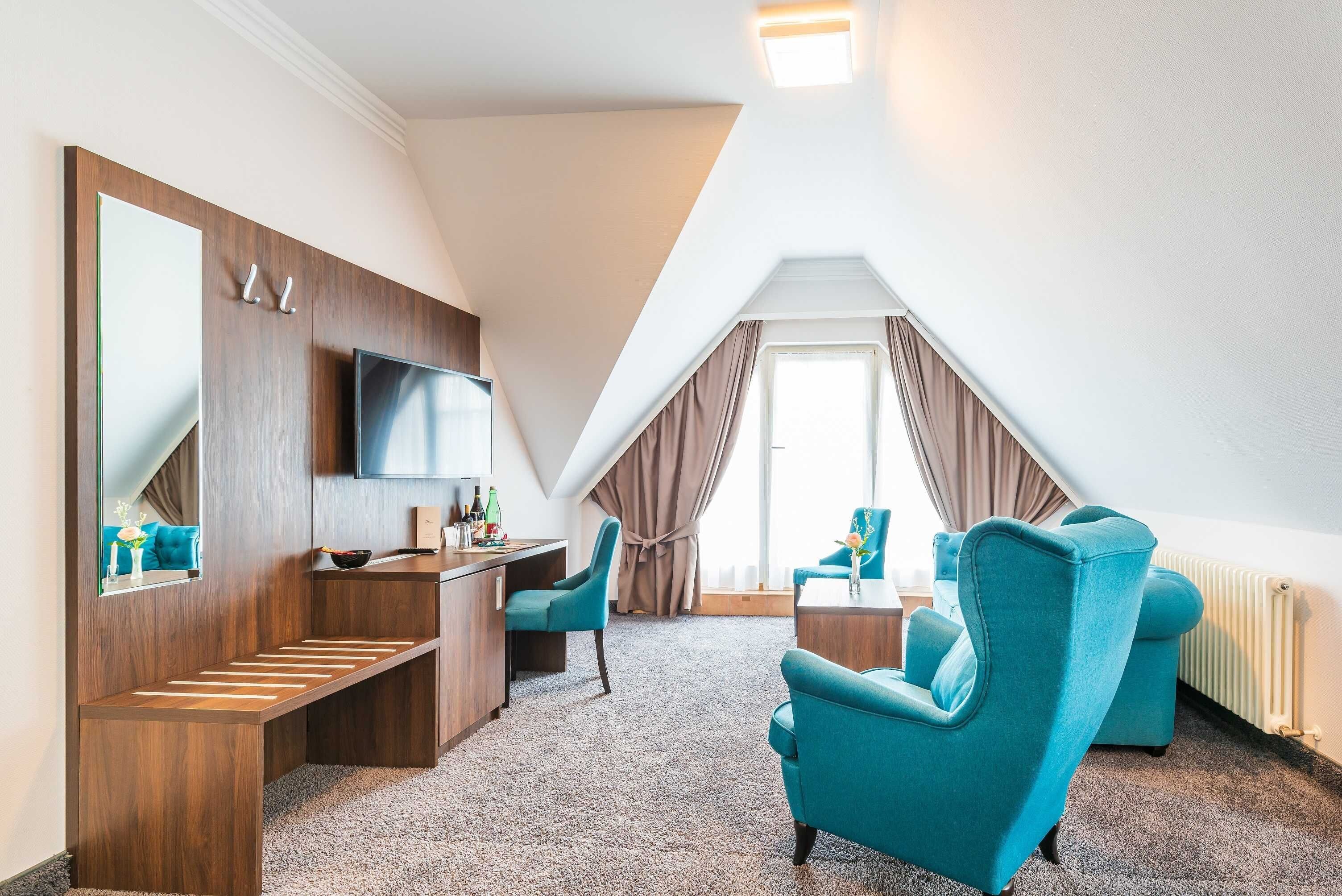 Ein modernes Hotelzimmer mit Schreibtisch aus Holz, Fernseher, großem Spiegel und zwei stahlblauen Sesseln.