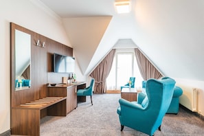 A modern hotel room with a wooden desk, a television, a large mirror, and two teal armchairs. - Junior Suite with Balcony - Hotel Villa Belvedere (Binz)
