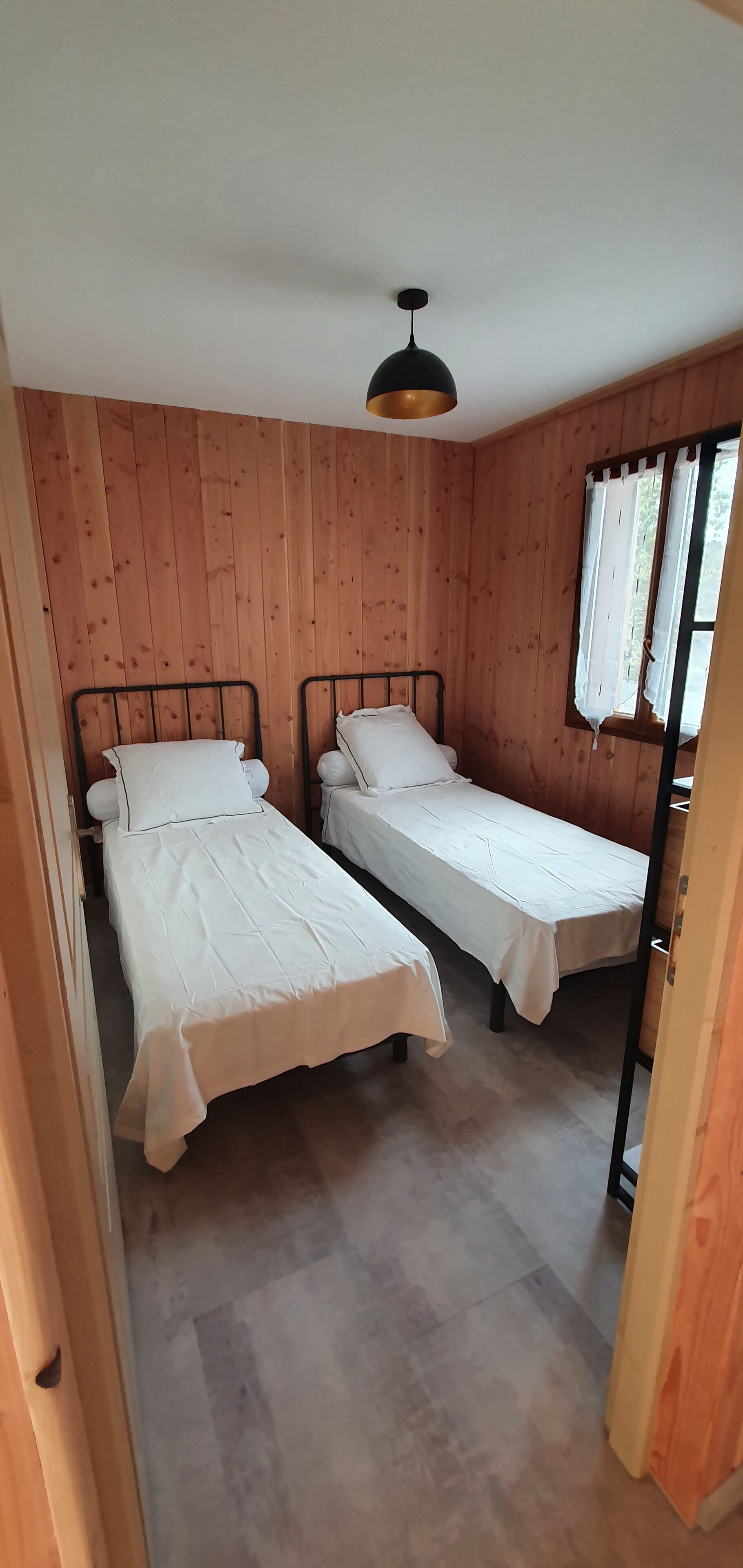 A room with two single beds, wooden walls, a ceiling light, and a window with curtains.