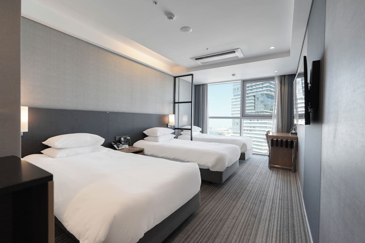 A hotel room with two beds, a large window with a city view, a flat-screen TV, and a minibar.