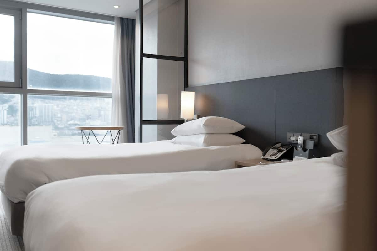 A hotel room with two beds, a bedside table, a lamp, a phone, and a window with a city view.