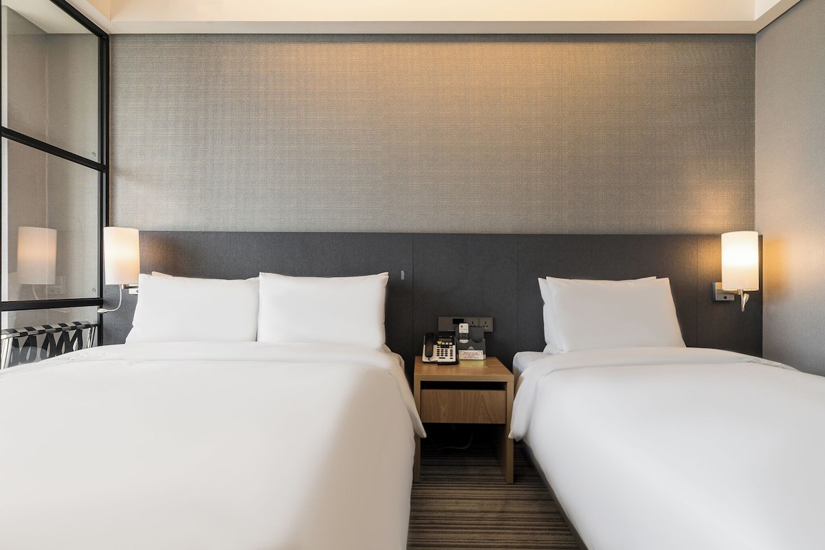 A hotel room with two beds, a nightstand with a phone, and wall-mounted lamps.