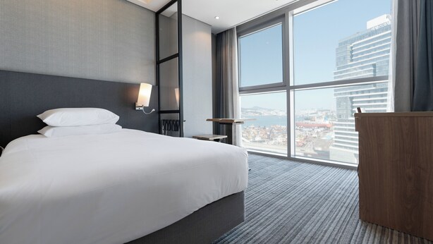 Una habitación de hotel moderna con una cama grande, un escritorio y vistas a un paisaje urbano a través de un amplio ventanal.