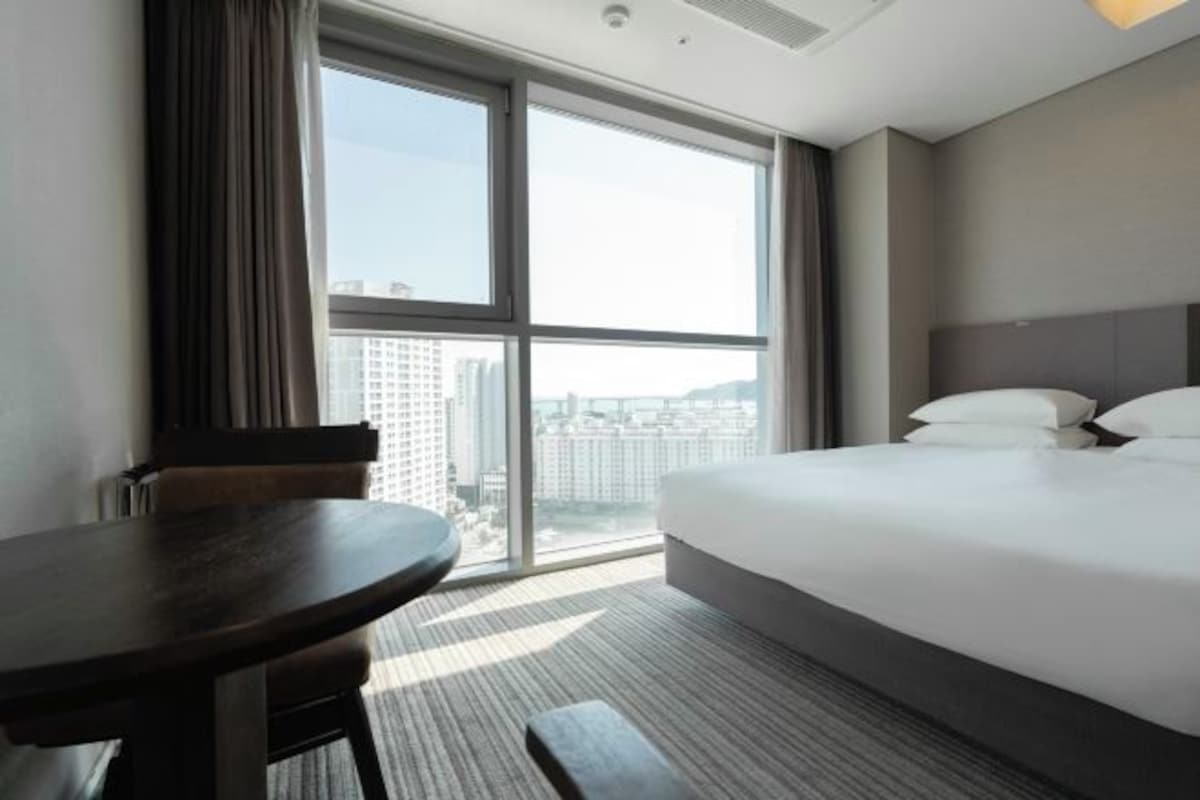 A hotel room with a large bed, a small table, and a view of the city through large windows.
