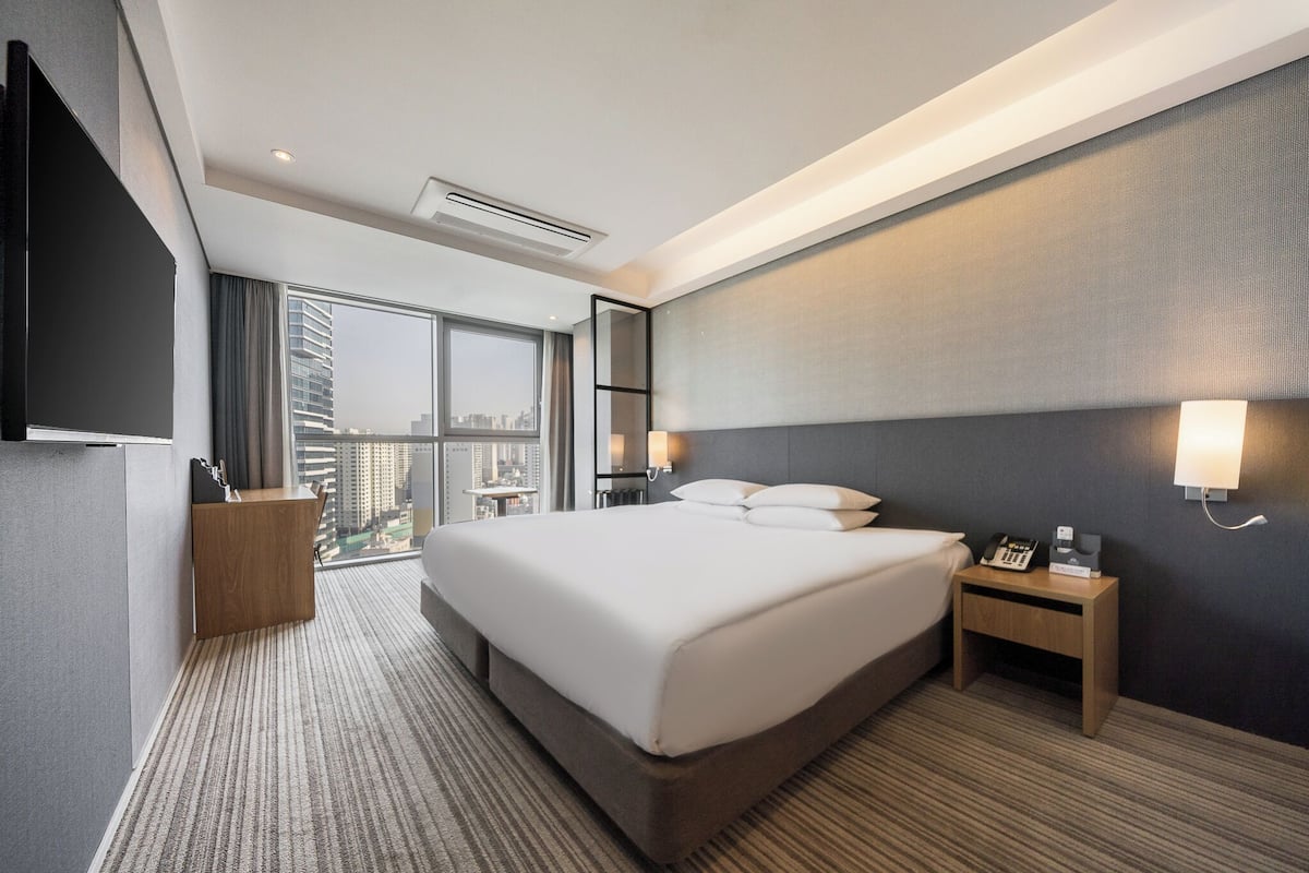 A hotel room with a large bed, a desk, a chair, a TV, a window with a city view, and a wall-mounted lamp.