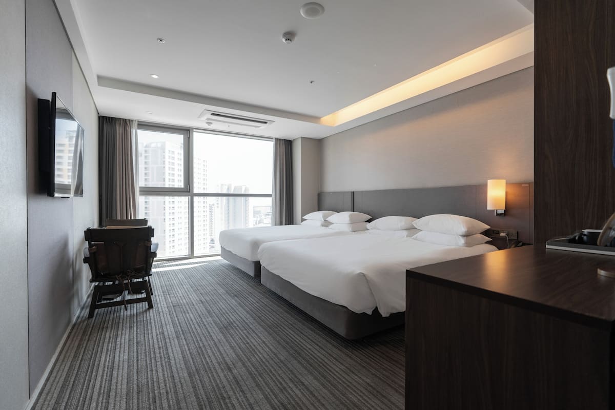 A hotel room with two beds, a desk, a chair, a TV, and a window with a view of buildings.