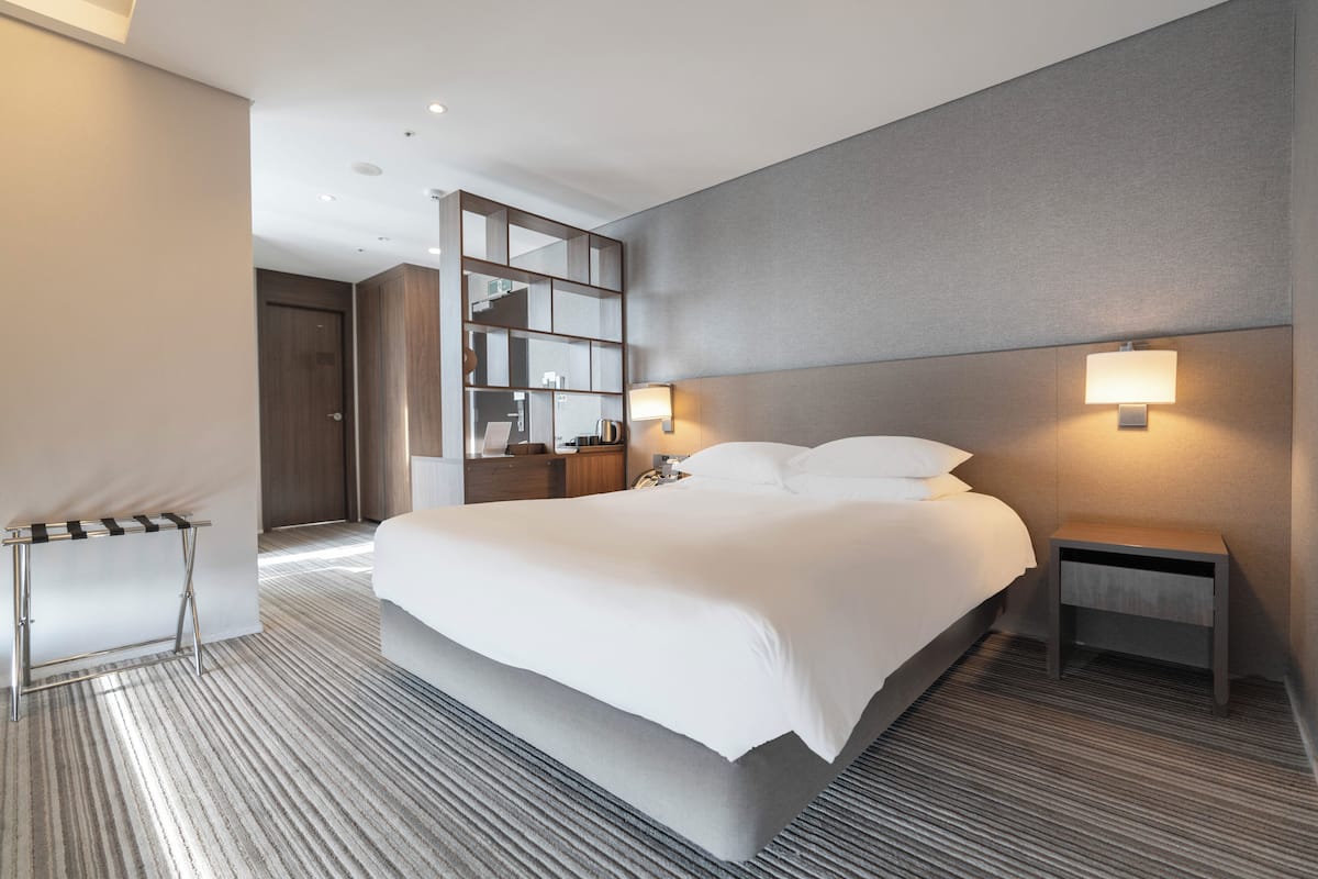 A modern hotel room with a large bed, a bedside table, a nightstand, a mirror, and a wall-mounted lamp.