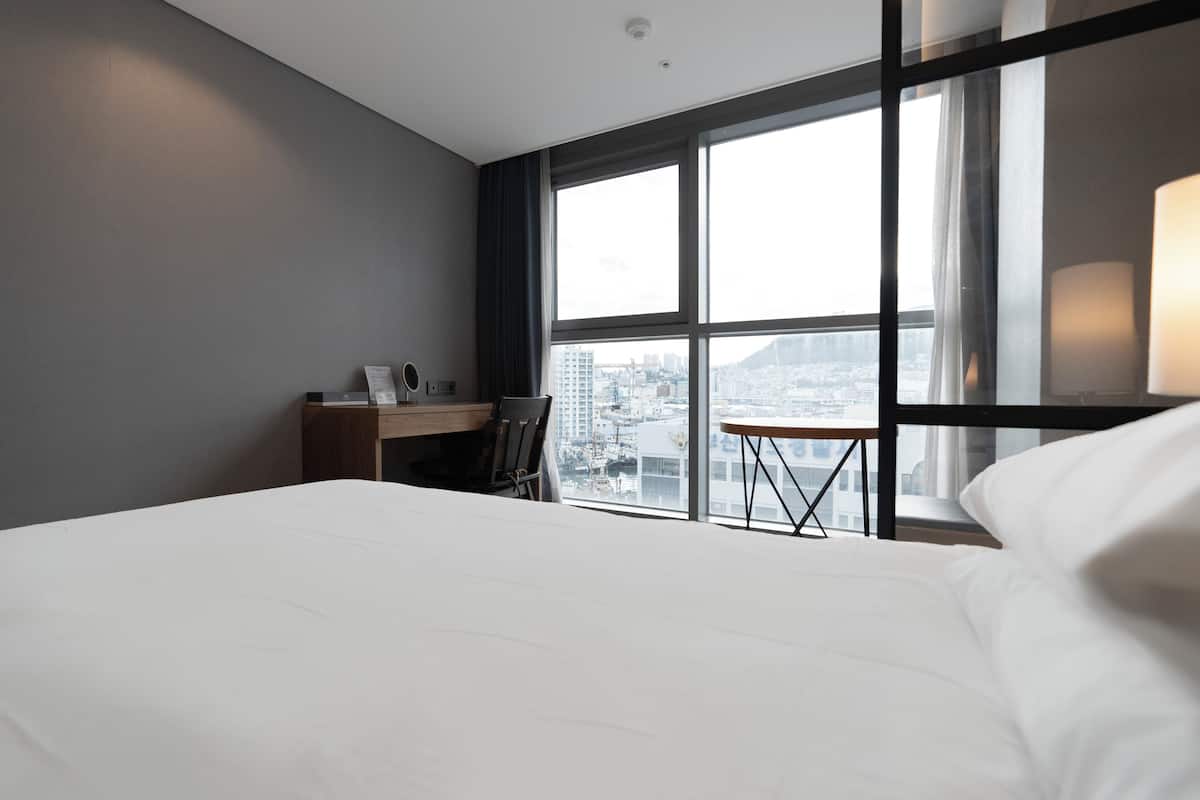 A hotel room with a large bed, a desk, and a chair. There is a window with a city view.