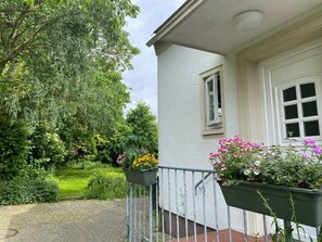 Ein Haus mit weißer Fassade, einem kleinen Fenster und einer Tür. Vor der Veranda stehen zwei Blumenkästen mit verschiedenen Blumen.