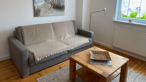 A living room with a grey sofa, a wooden coffee table with a woven basket, a floor lamp, and a window with a view of greenery. - Apartment Heimsoth Kappeln EG - Apartments Heimsoth in Kappeln (Kappeln)