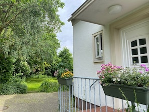 Ein Haus mit weißer Fassade, einem kleinen Fenster und einer Tür. Vor der Veranda stehen zwei Blumenkästen mit verschiedenen Blumen.