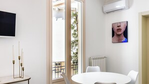 A room with a round table, white chairs, a TV mounted on the wall, and a window with a view of a balcony. - Alma Suite - Two Bedroom Apartment, Sleeps 6 (Noto)