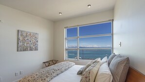 A bedroom with a large window, a bed with patterned bedding, a small table, and a painting on the wall.