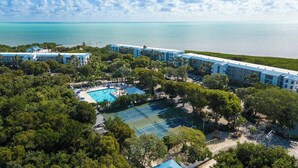 A resort with multiple buildings, a swimming pool, tennis courts, and a view of the ocean.