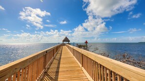 A wooden pier leading to a thatched-roof structure with a view of the sea and sky.