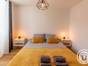 Un dormitorio con cama, dos mesitas de noche, una lámpara de techo con diseño geométrico y un logotipo en la pared.