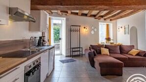 A modern kitchen with a built-in oven, a coffee machine, and a sink. There is a dining area with a brown sofa and a table. The room has a wooden ceiling and a view of the outdoors. - Charming Riverside Home in UNESCO Heritage Village with Modern Amenities & Near Angers (Bouchemaine)