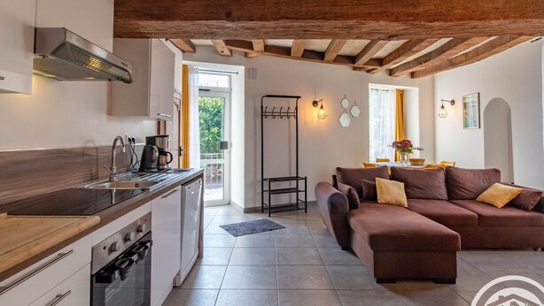 Eine moderne Küche mit Einbauofen, Kaffeemaschine und Spüle. Es gibt eine Essecke mit einer braunen Couch und einem Tisch. Der Raum verfügt über eine Holzbalkendecke und einen Blick ins Freie.
