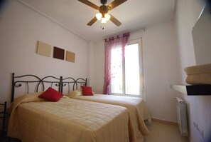 Una habitación con dos camas, ventilador de techo, ventana con cortinas y radiador.