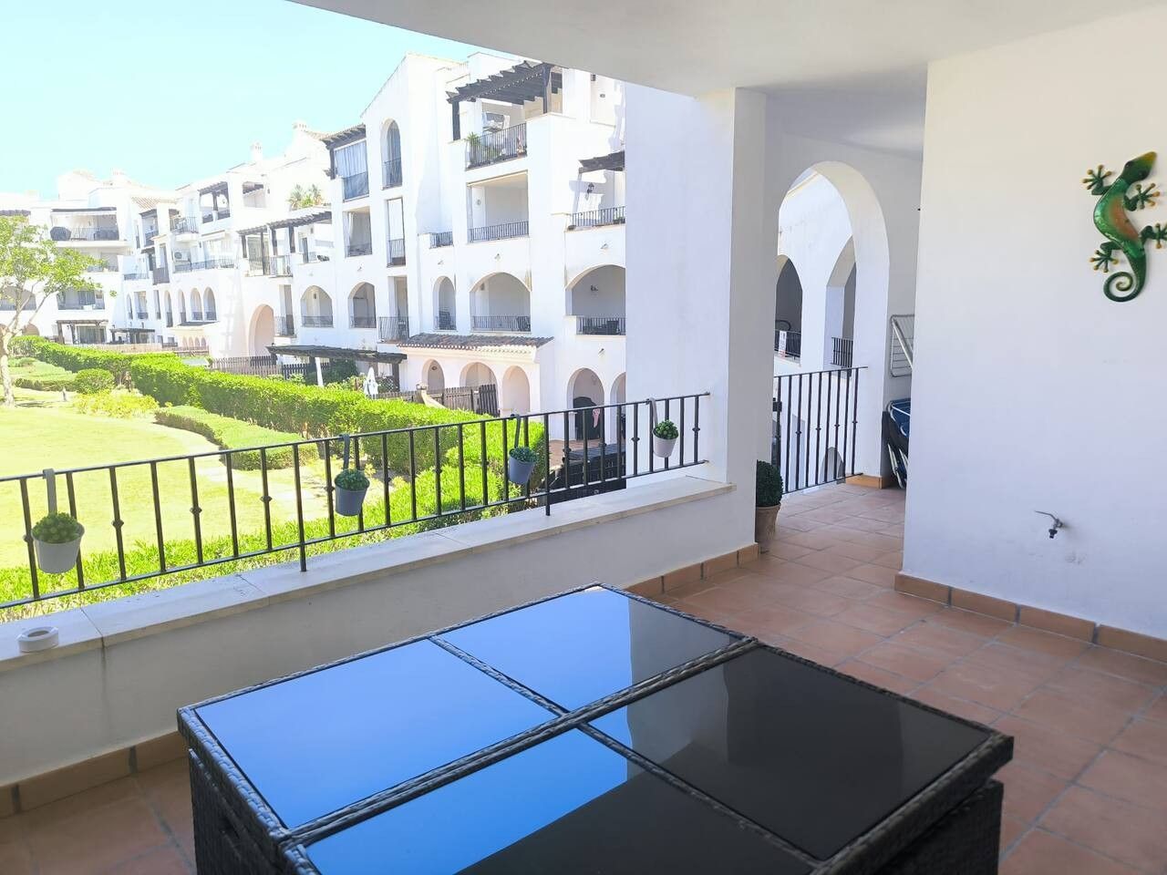 A balcony with a glass table, white walls, and a view of a courtyard with buildings.