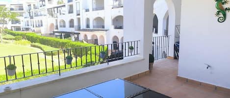 A balcony with a glass table, white walls, and a view of a courtyard with buildings.