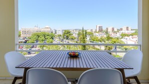 En balkong med ett träbord och två vita stolar, med utsikt över en stadsmiljö med byggnader och träd.