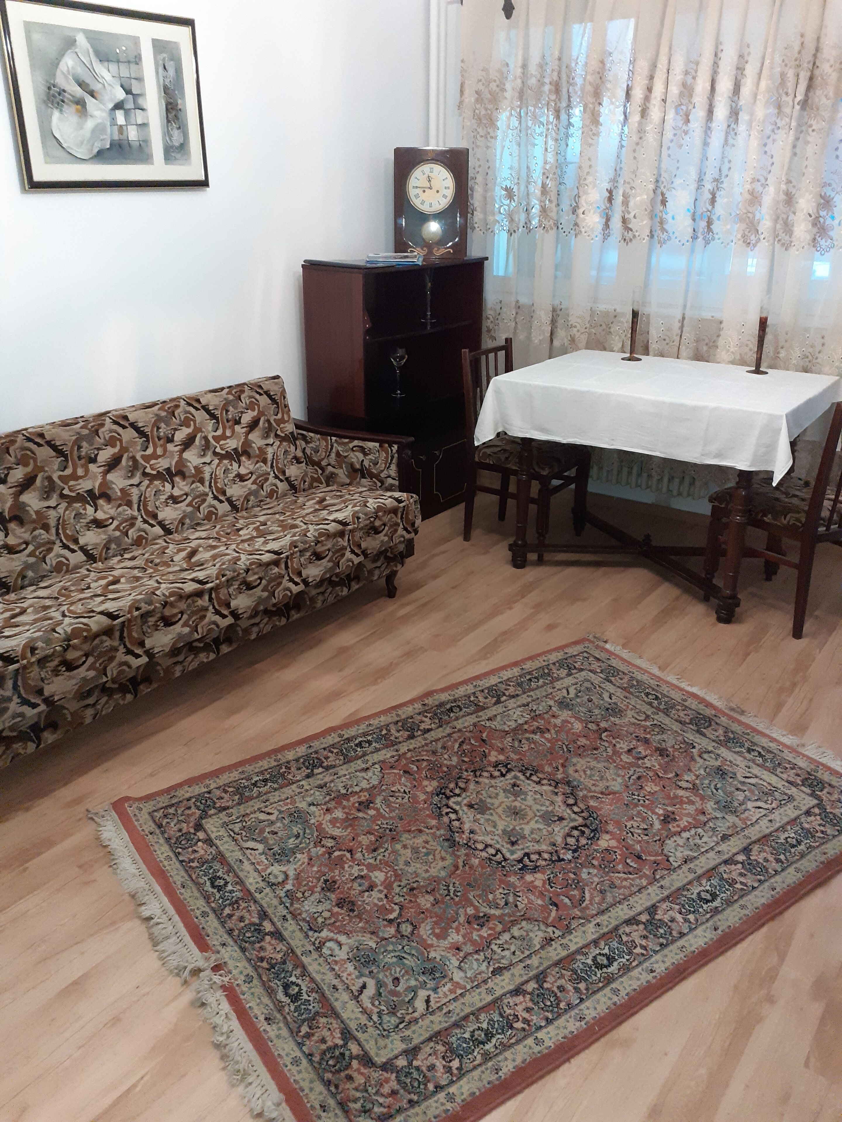 A living room with a patterned sofa, a dining table with chairs, a wooden cabinet, a clock, and a large rug.