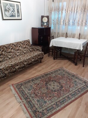 Uma sala de estar com um sofá estampado, uma mesa de jantar com cadeiras, um armário de madeira, um relógio e um tapete grande.